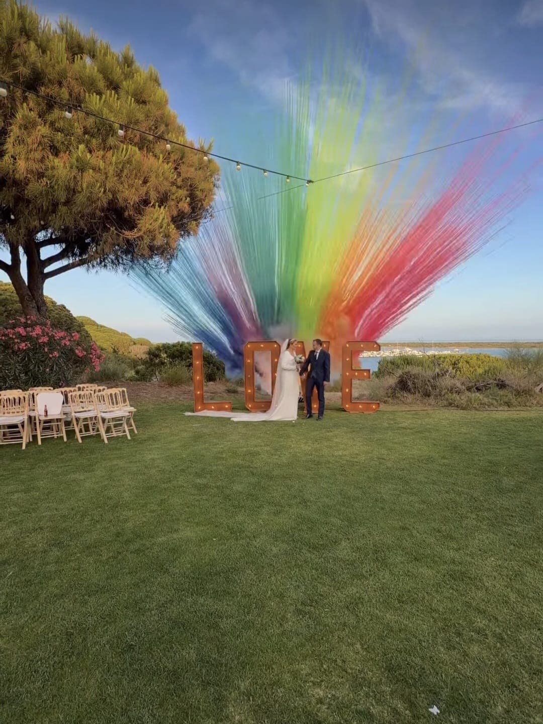 Noivos junto às letras LOVE, com um leque de fumos coloridos num casamento